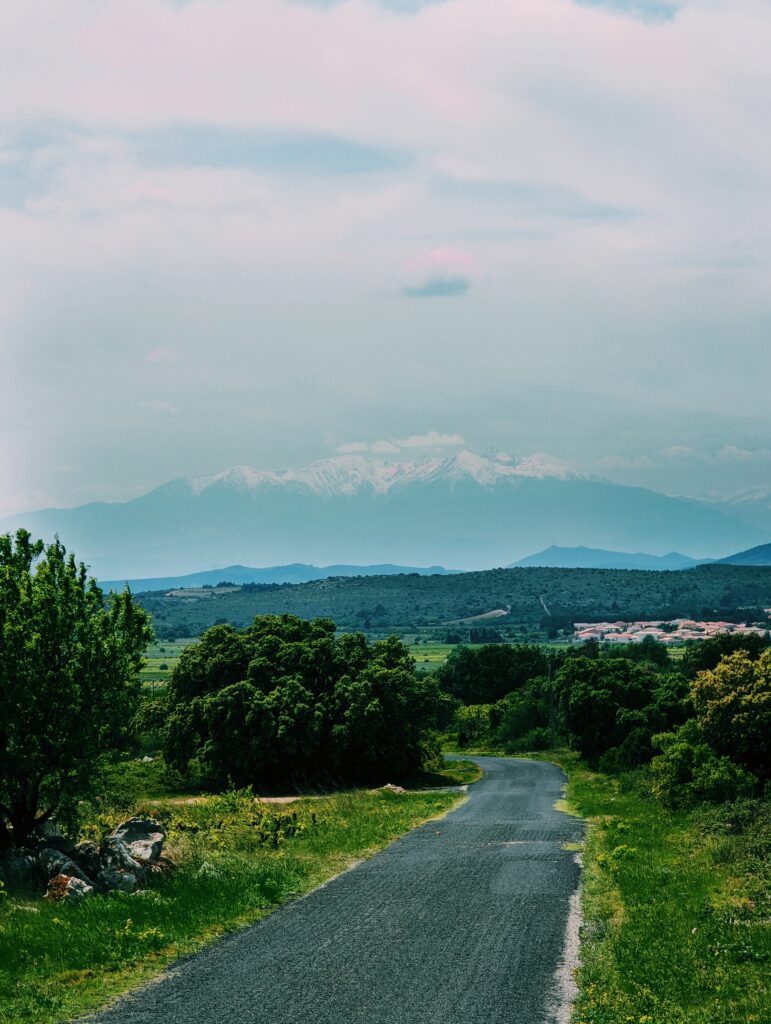 A road winding down the hill in the foreground, view over Roussillan plains and Pic du Canigou in the background