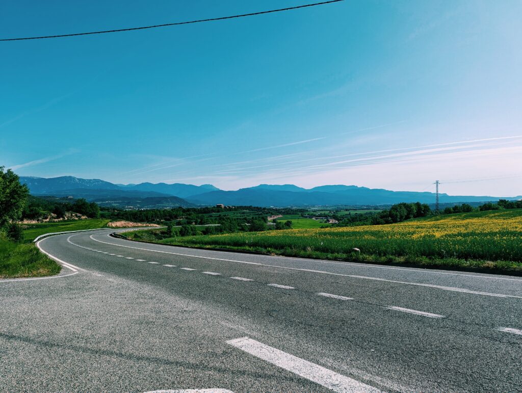 Looking northeast back on the foothills of the Pyrenees. A road winding up a hill in the foreground with some houses in the middleground. Bright sun and clear blue skies.