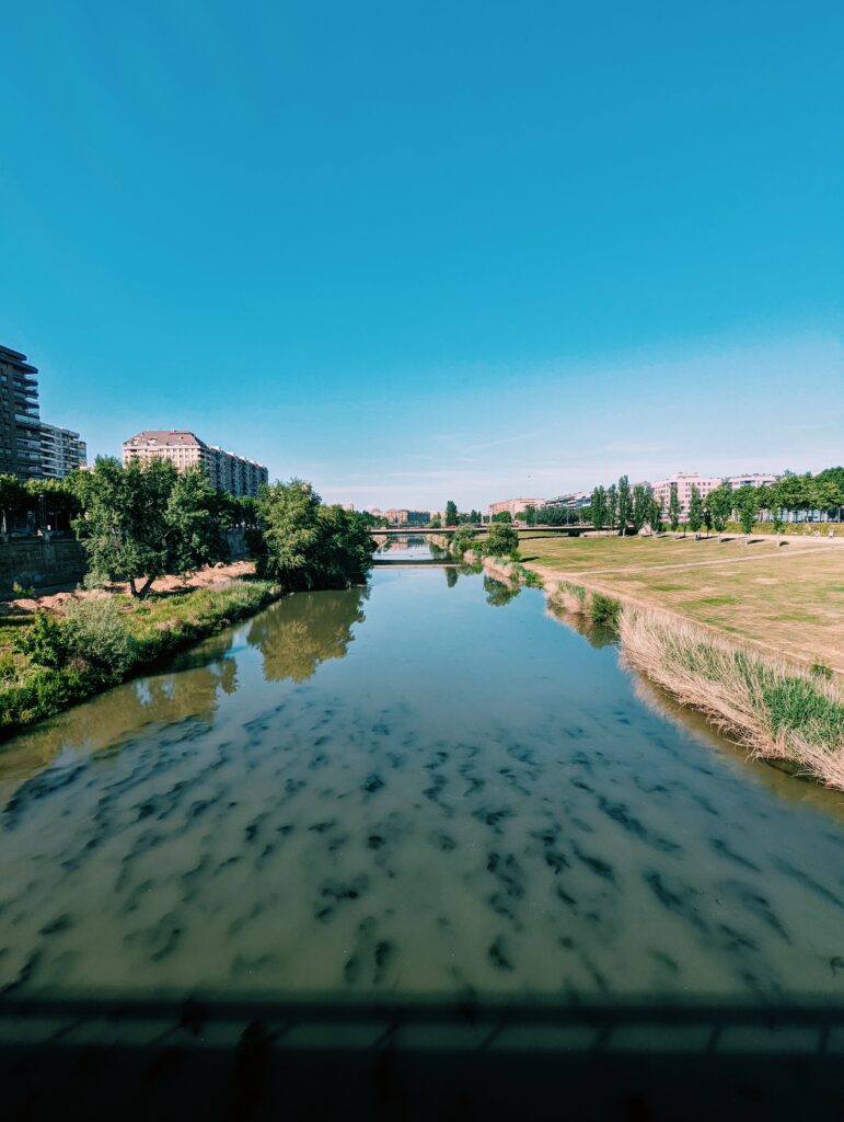 Rio Segre in Lleida, taken from a bridge in the middle of the river trees and buildings can be seen on both banks and another bridge in the distance