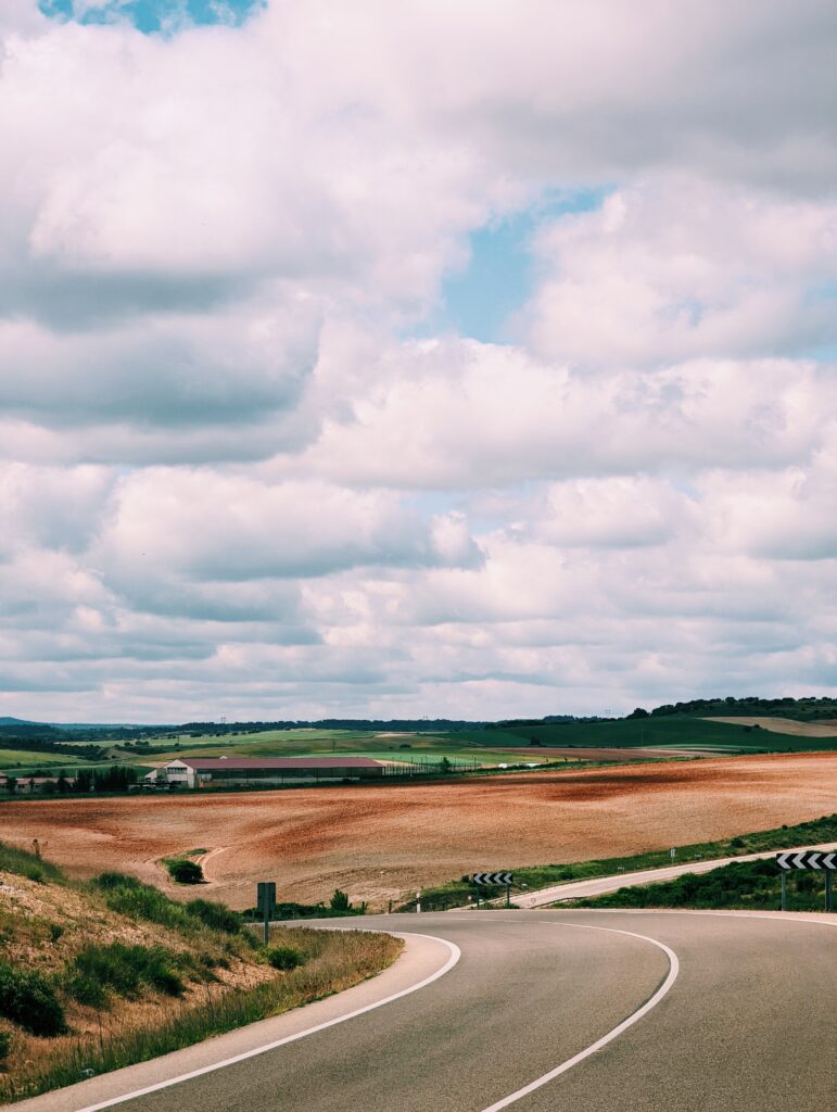 Road in the foreground winding down a hill, fields in the middleground and mostly cloudy skies.